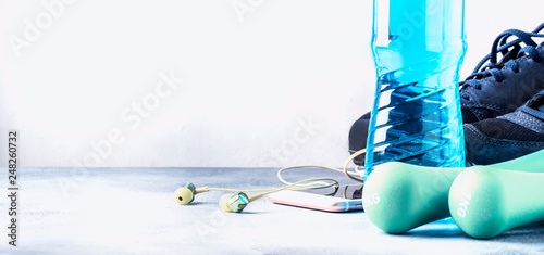 Female fitness concept: light dumbbells, isotonic blue water, smartphone with headphones and running shoes on gray concrete floor, selective focus