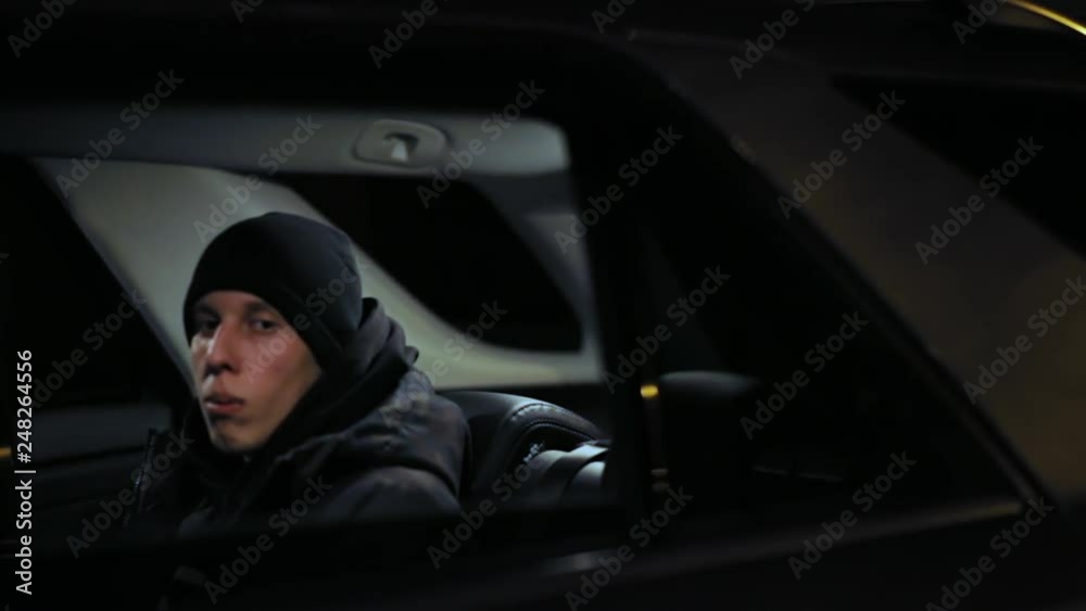 Guy in black hat sitting at backseat of car, looking through window, swaying to rhythm. Night time. Criminal concept. Thief sitting in car, steal, tool Guy in black hat sitting at backseat of car, looking through window, swaying to rhythm. Night time. Criminal concept. Thief sitting in car, steal, tool