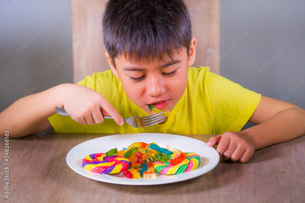 young happy and excited male kid smiling cheerful eating dish full of candy and lollipop sitting at table isolated on grey background in sugar addiction and abuse young happy and excited male kid smiling cheerful eating dish full of candy and lollipop sitting at table isolated on grey background in sugar addiction and abuse