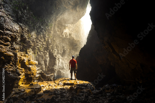 Portrait of a Male Backpacker wearing a red shirt standing in sunlight in a cave beneath the Elephant waterfall in Dalat Vietnam.