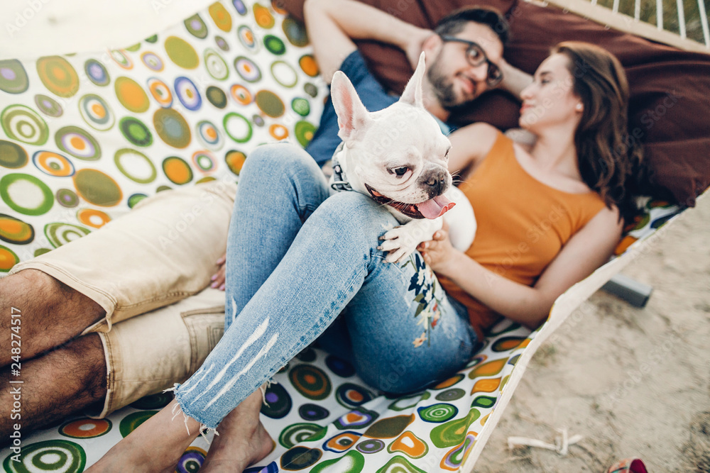 Obraz premium happy hipster couple with bulldog relaxing in hammock on the beach in sunset light, summer vacation. stylish family with dog cuddling and having fun, cute moments in summer evening
