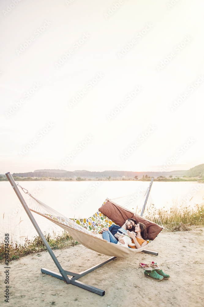 happy hipster couple with bulldog relaxing in hammock on the beach in sunset light, summer vacation. stylish family with dog cuddling and having fun, cute moments in summer evening