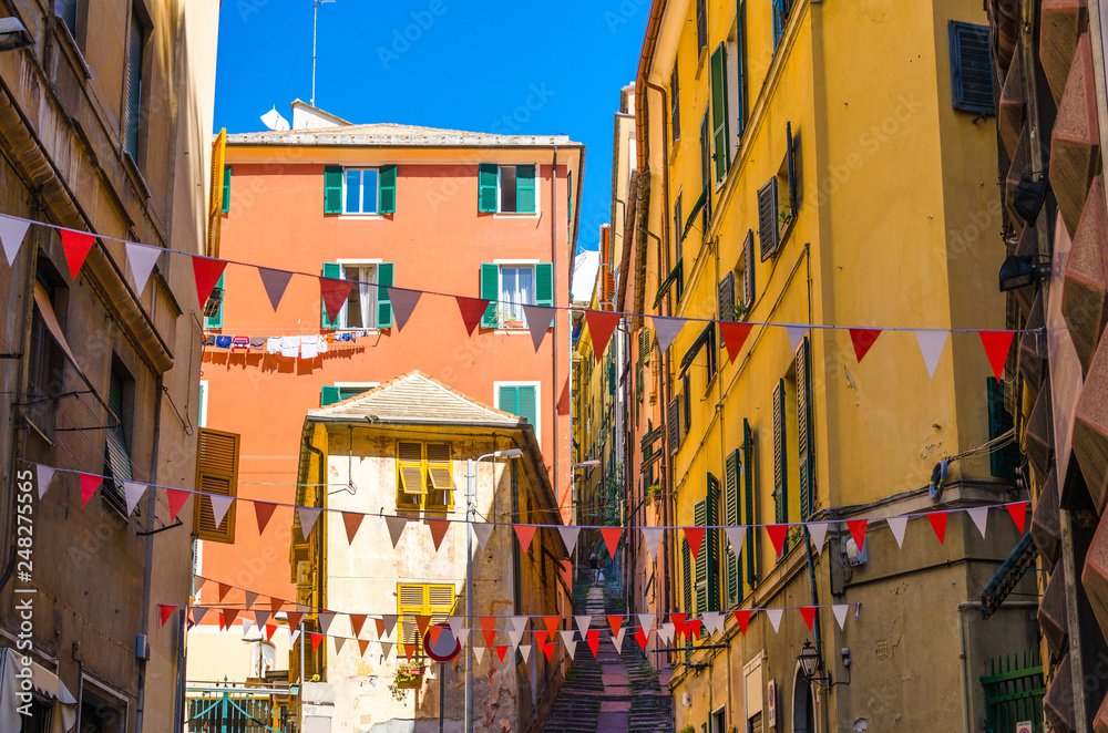 Small red and white flags hang above street with colorful multicolored buildings with shutters on windows in historical centre of old european city Genoa (Genova), Liguria, Italy