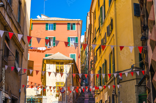 Small red and white flags hang above street with colorful multicolored buildings with shutters on windows in historical centre of old european city Genoa (Genova), Liguria, Italy