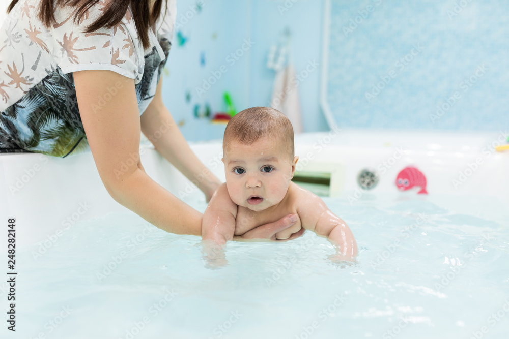 Newborn baby swimming in the bath or bathtub. Mother help him swim. Mom