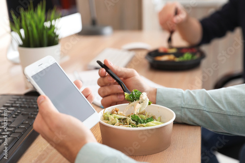 Office employee with smartphone having lunch at workplace, closeup. Food delivery photo