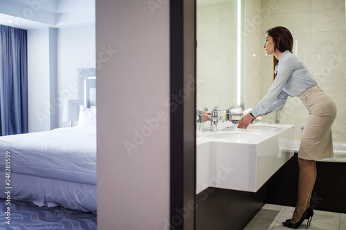 Business lady in dressed in button-down shirt, pencil skirt and stilettos, looking at her reflection in the mirror while washing hands in the bathroom