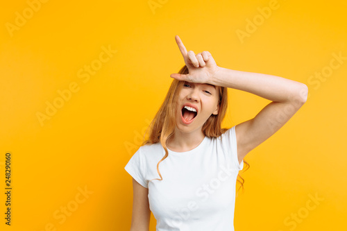 girl shows the gesture of a loser or loser, in a white t-shirt with a grin on his face, on a yellow background photo