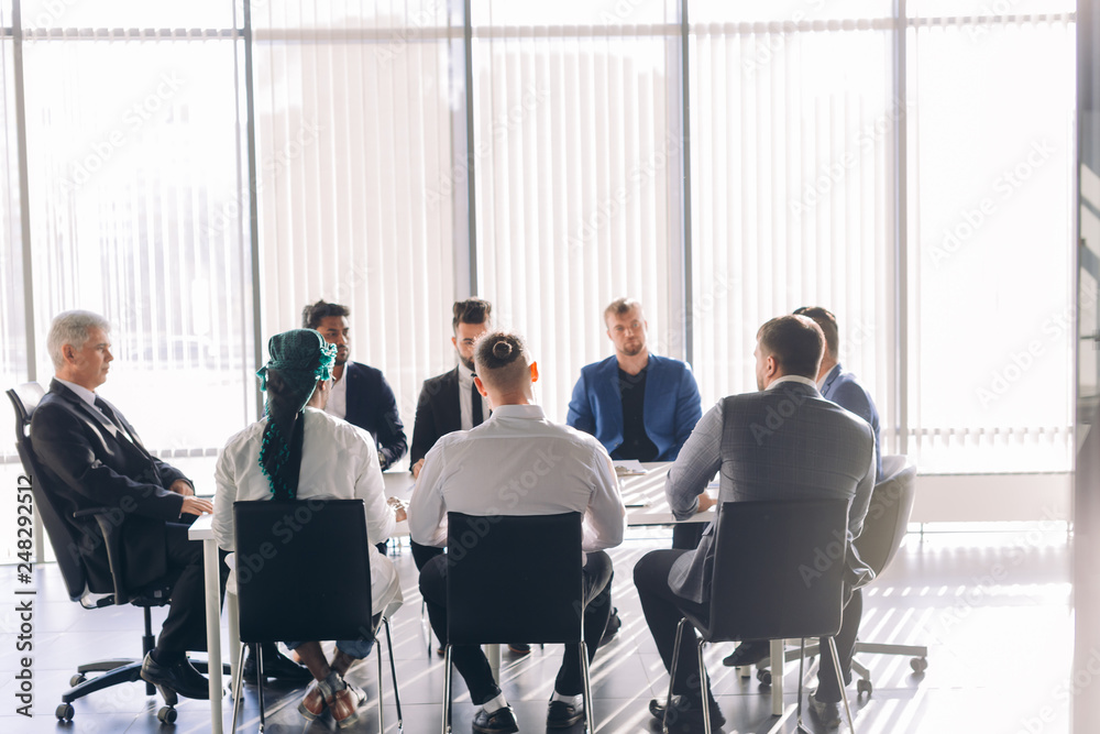 Multiracial male business executives discussing project sitting at conference table in spacious hall with panoramic city scapes, all men dressed in formalwear and have positive mood