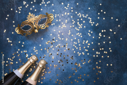 Two champagne bottles, golden carnival mask and confetti stars  on blue background. Christmas background, top view, copy space.