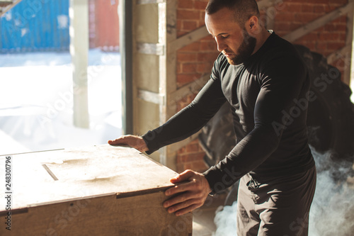 Handsome strong caucasian bearded man prepares to jumps onto a plyobox in a cross training gym with big panoramic windows and giant tire on background photo