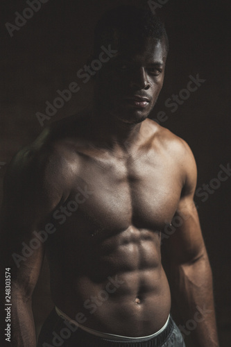 Attractive African male fighter or boxer posing shirtless, looking at camera, isolated over dark background.