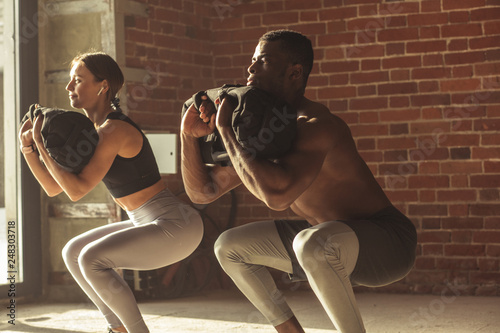 Obraz na plátně Young mixed-race fit couple doing squats exercise, with heavy sandbag in gym