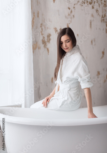 Portrait of happy beautiful woman with brown long hair, sitting near bath tube in bathroom with modern interior, healthy skin and beauty of youth. Fresh clean stylish wellness beauty lifestyle.