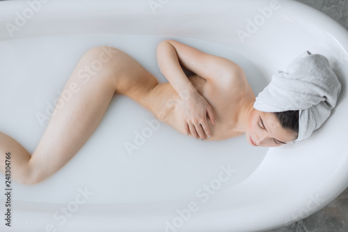 High angle view of cute caucasian female model with perfect slim figure lying in milky water in seductive pose with towel on head. Perfect healthy Skin. Skincare. Spa salon.