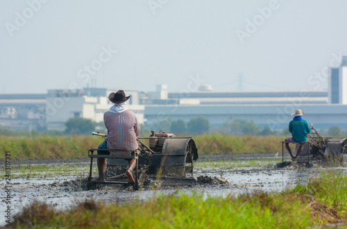 Thai farmer driving tiller tractor to plow paddy field prepare new rice with industrial factory background.