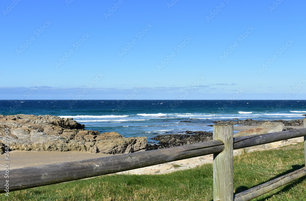 Beach with grass, wooden fence and morning light. Rocks and blue sea with waves, sunny day. Galicia, Coruña Province, Spain.