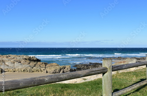 Beach with grass  wooden fence and morning light. Rocks and blue sea with waves  sunny day. Galicia  Coru  a Province  Spain.