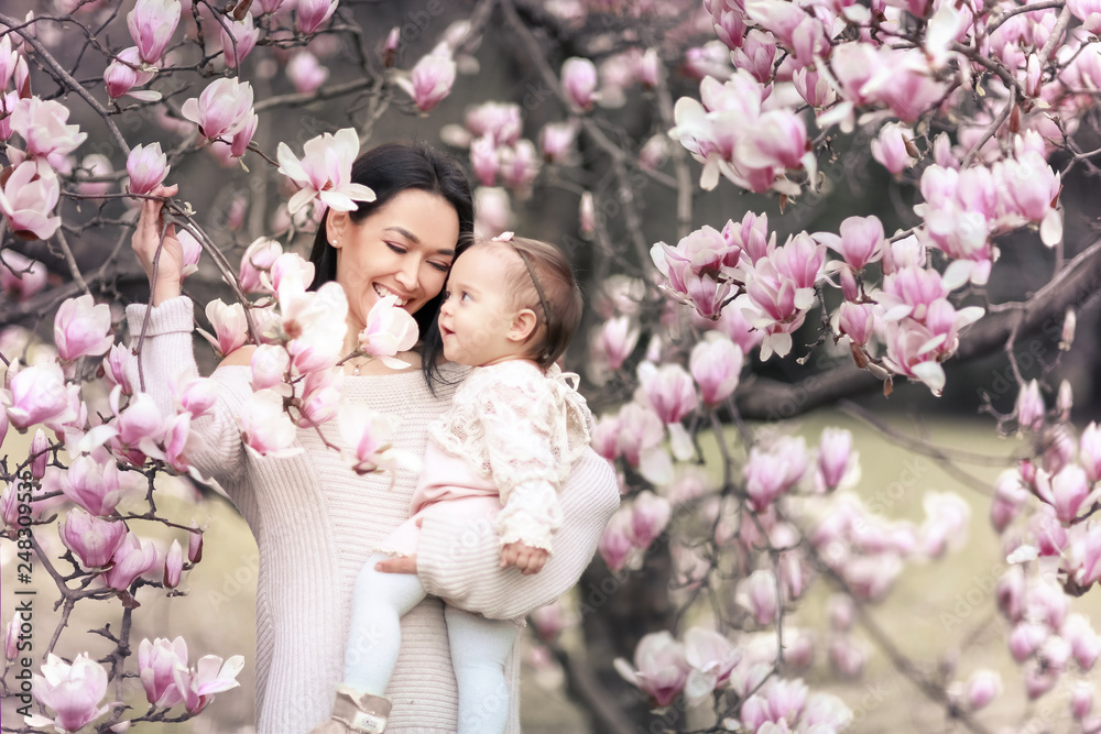 Obraz premium Cute Baby 6 month old Girl in Pink Outfit with Big Blue Eyes with Young Beautiful Mother at Spring, Pink Blooming Tree at the Background