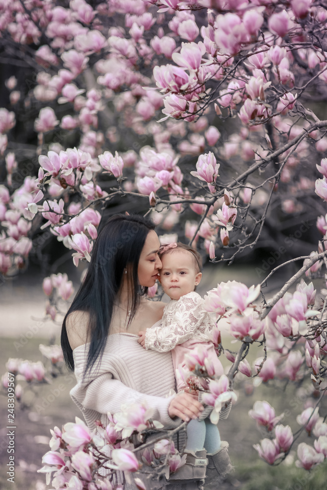 Fototapeta premium Cute Baby 6 month old Girl in Pink Outfit with Big Blue Eyes with Young Beautiful Mother at Spring, Pink Blooming Tree at the Background