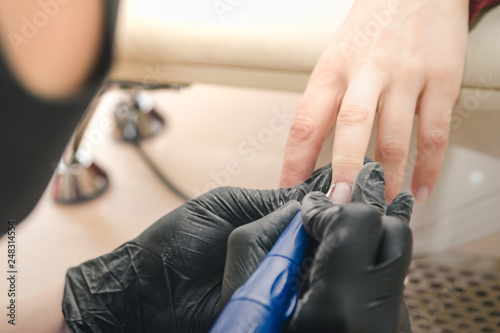 A manicurist in black latex gloves makes a hardware manicure to a client using a manual electric milling machine in a beauty salon.