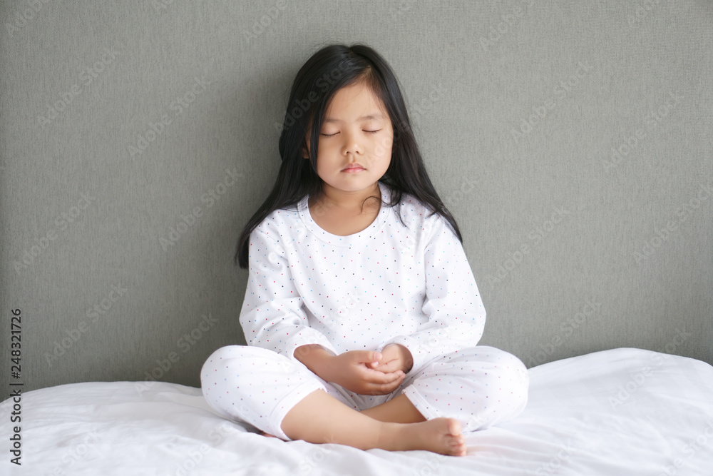 Asian child cute or kid girl sit meditation with peace or quiet and relax for mindfulness and concentrated breath on wake up in morning on bedroom at home and wear white dress on gray background Asian child cute or kid girl sit meditation with peace or quiet and relax for mindfulness and concentrated breath on wake up in morning on bedroom at home and wear white dress on gray background