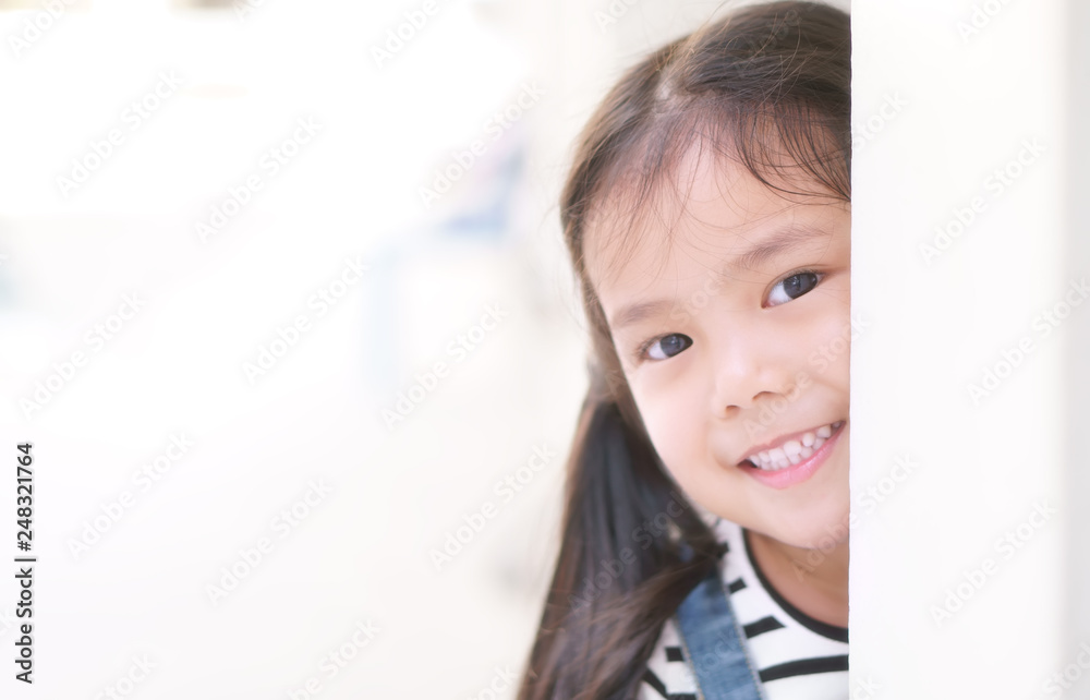 Asian child curious or kid girl playing peekaboo or hide and seek with sneaked or dodge behind post and happy smiling white teeth or cheerful at public park or playground on relax holiday with space