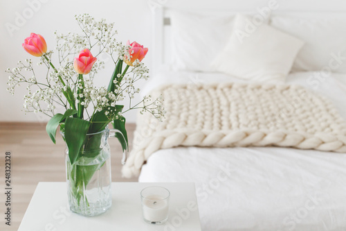 Glass vase with pink tulips and gypsophila flowers in light cozy bedroom interior. White wall, bed with white linen, beige thick yarn knitted woolen merino chunky blanket or plaid, pillows, candle.