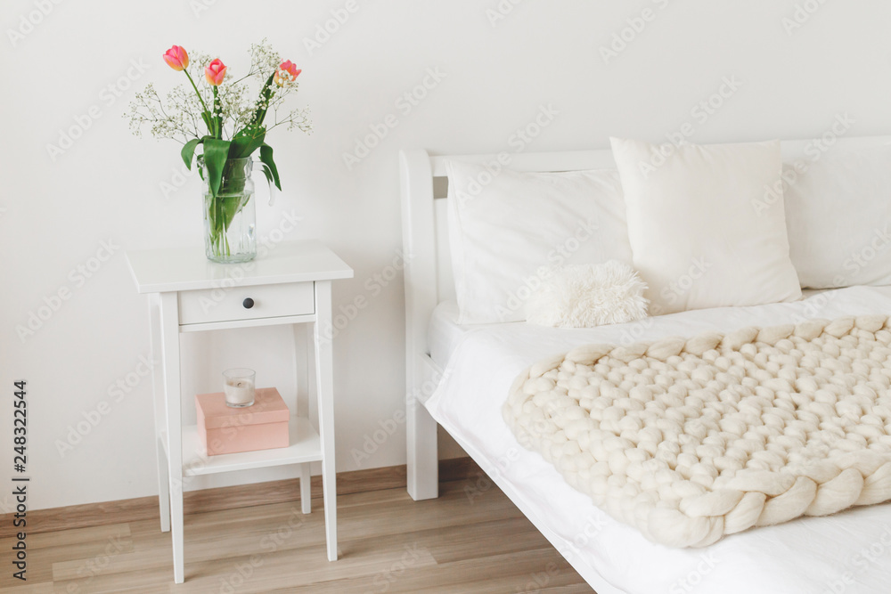 Naklejka premium Cozy bedroom interior: white wall, bed with white linen, light beige thick yarn knitted woolen merino chunky blanket or plaid, pillows, bedside table, vase with tulips flowers.