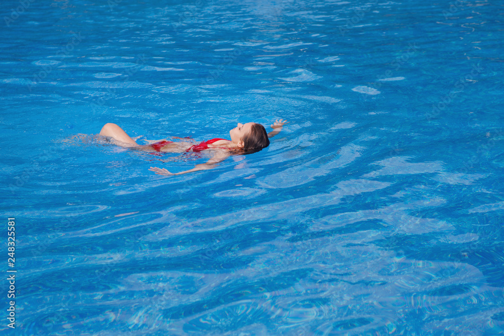 Young cute woman with long hair in a red bikini swims on her back in a pool with blue water in a resort hotel.