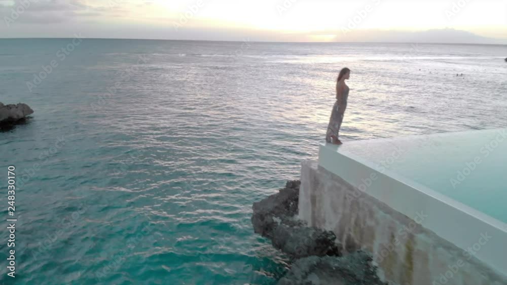 Aerial: Woman Standing on the Edge of Swimming Pool next to the Ocean at Sunset