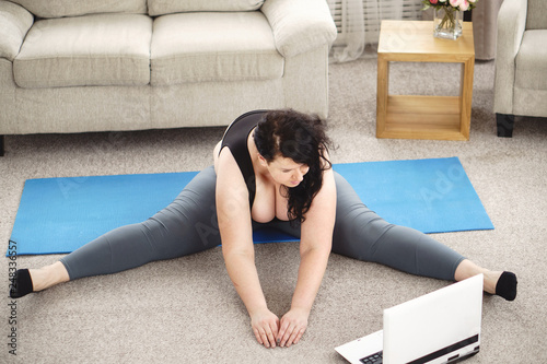 fitness, online personal trainer, home training, technology and diet. overweight woman doing stretching exercises sitting on a fitness mat at home, using laptop