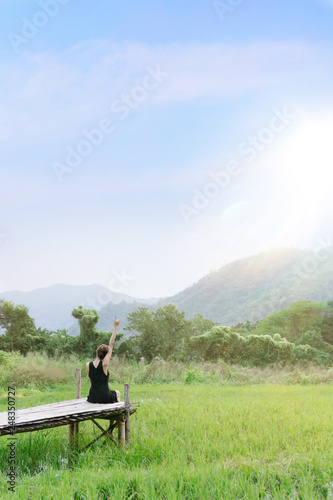 A woman with black dress sitting on a the wooden bridge and raise her hand and making the sign “I love you” on the fresh green field with mountains, blue sky and sunlight flare.