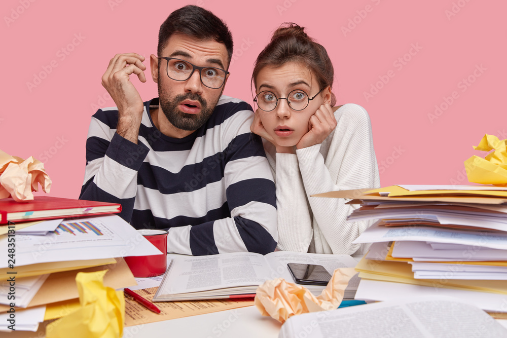 Indoor shot of surprised coworkes have astonished facial expressions, wear transparent glasses, cramm material for final examination, pose over pink background. People and partnership concept Indoor shot of surprised coworkes have astonished facial expressions, wear transparent glasses, cramm material for final examination, pose over pink background. People and partnership concept