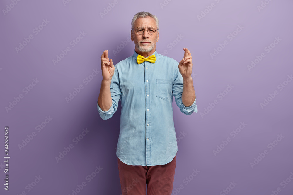 Hopeful intense male pensioner keeps fingers crossed, makes wish and puts efforts to pray, wears yellow bowtie, denim shirt and trousers, models over purple studio background. Dreams come true Hopeful intense male pensioner keeps fingers crossed, makes wish and puts efforts to pray, wears yellow bowtie, denim shirt and trousers, models over purple studio background. Dreams come true
