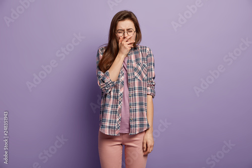 Sad woman in stress cries desperately, has embarrassed facial expression, expresses sadness, suffers from hurt feelings, dressed in checkered shirt, casual trousers, isolated over purple background