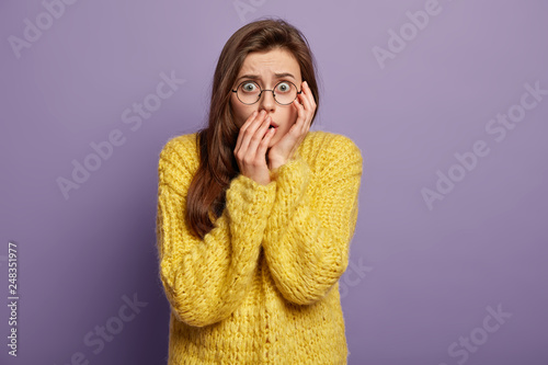 Waist up shot of fearful shocked stupefied young woman covers mouth, has eyes popped out, dark hair, dressed in loose knitted yellow sweater, isolated over purple studio background. Omg concept
