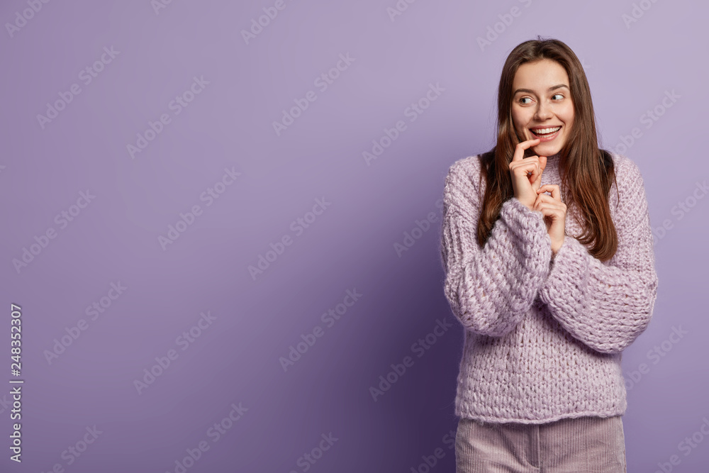 Flirty young girl in winter loose sweater smiles sensually, glances aside, keeps hand near mouth, rejoices something pleasant, focused aside, isolated over purple background with empty space left Flirty young girl in winter loose sweater smiles sensually, glances aside, keeps hand near mouth, rejoices something pleasant, focused aside, isolated over purple background with empty space left