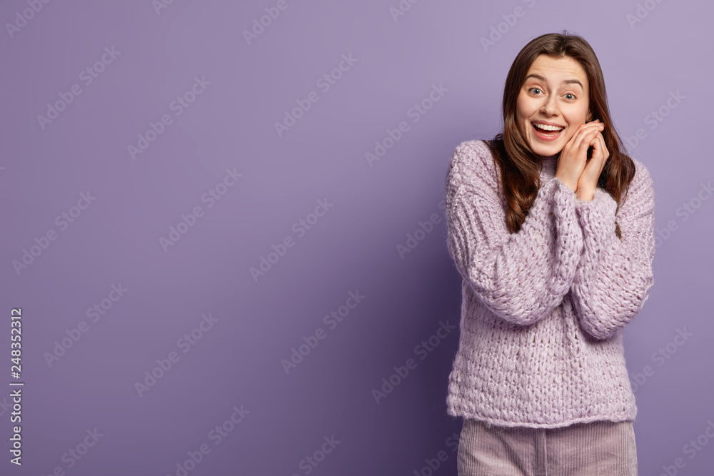 Indoor shot of happy positive woman smiles broadly, enjoys cold frosty winter weather, wears warm knitted clothes, keeps hands near face, models over lilac studio wall with empty space aside Indoor shot of happy positive woman smiles broadly, enjoys cold frosty winter weather, wears warm knitted clothes, keeps hands near face, models over lilac studio wall with empty space aside