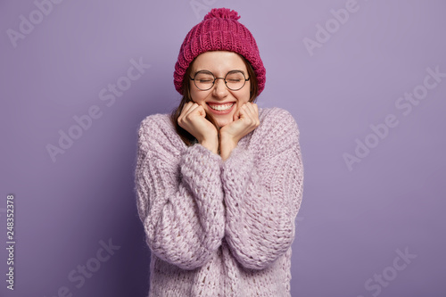 Photo of happy European girl hears positive news, keeps hands under chin, wears hat and jumper, smiles broadly, rejoices good winter day, isolated over purple background. Cheering energetic woman photo