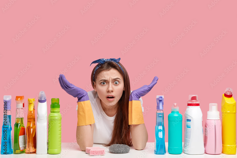 Isolated shot of puzzled hesitant young woman spreads palms, has displeased expression, wears stylish headband, uses cleaning detergents, isolated over pink background with empty space above Isolated shot of puzzled hesitant young woman spreads palms, has displeased expression, wears stylish headband, uses cleaning detergents, isolated over pink background with empty space above