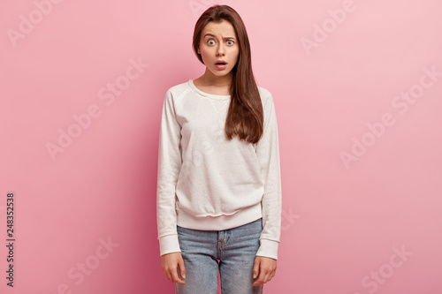 Studio shot of puzzled human raises eyebrows, has scared facial expression, dressed in white sweatshirt and jeans, isolated over rosy background. Pretty woman expresses disbelief, opens mouth