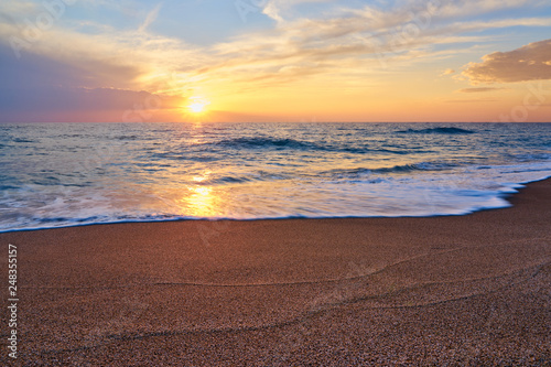 Colorful sunset at the tropical beach, sun behind clouds reflects on water and waves with foam hitting sand.