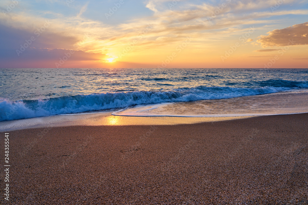 Colorful sunset at the tropical beach, sun behind clouds reflects on water and waves with foam hitting sand.