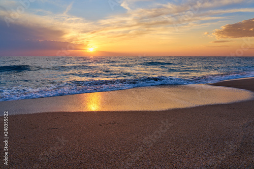 Colorful sunset at the tropical beach, sun behind clouds reflects on water and waves with foam hitting sand. © Arthur