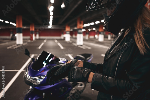 Cropped sideways shot of blonde female rider in black jacket and safety helmet putting on leather gloves, preparing for ride on stylish chopper, standing in deserted underground parking lot