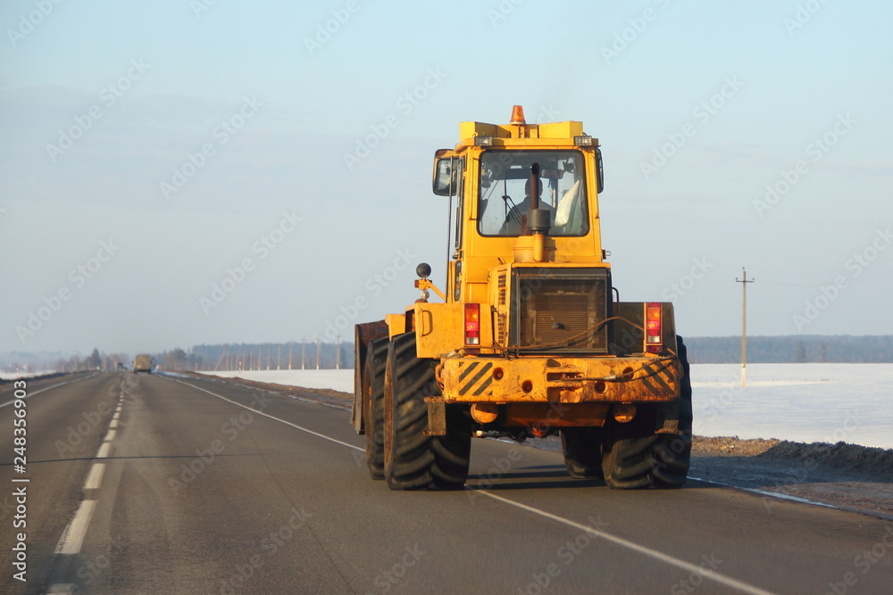 Heavy yellow bulldozer tractor rides along the asphalt road on the background of snowy fields and the blue of a clear sky in winter - back view