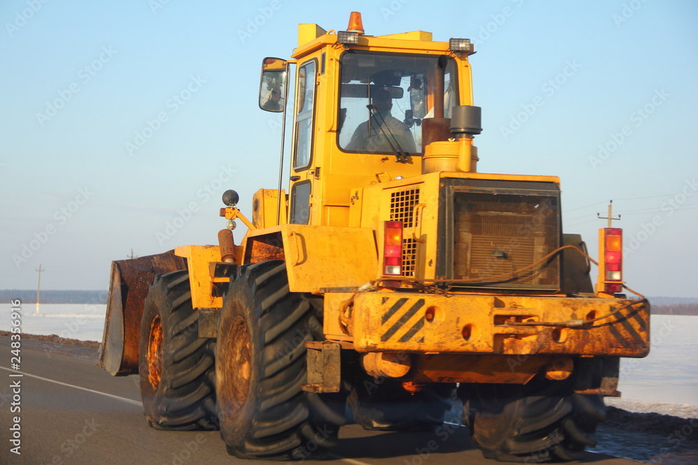 Large yellow bulldozer tractor rides along the asphalt road on the background of snowy fields and the blue of a clear sky in winter close up