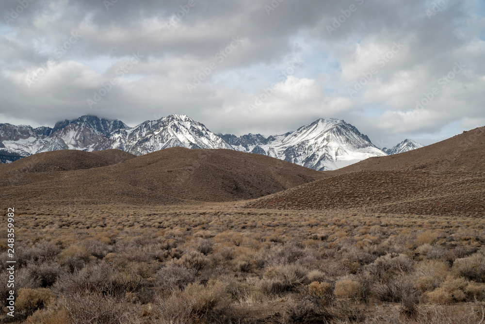 brown winter desert valley landscape with brown rolling hills, snowy mountain peaks of Sierra Nevada and gray cloudy sky in California, USA