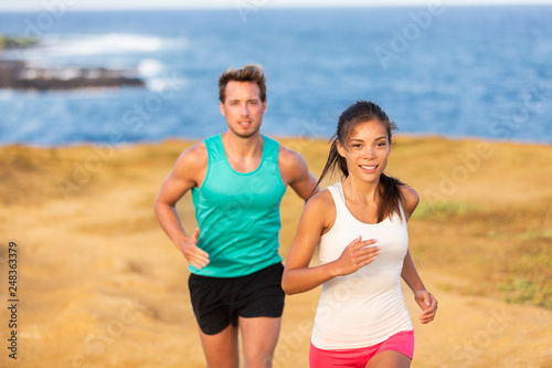 Fit run people couple jogging for fitness running on beach landscape nature outdoors. Woman and man sports athletes training cross-country trail running. Team partners, Asian woman, Caucasian man.